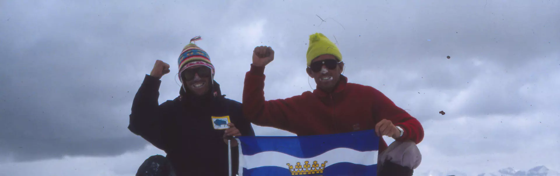 Twee mensen in winterkleding knielen op de besneeuwde top van een berg; ze glimlachen en steken een vuist in de lucht, terwijl ze een blauw-witte vlag met een kroon en leeuwen vasthouden. Op de achter
